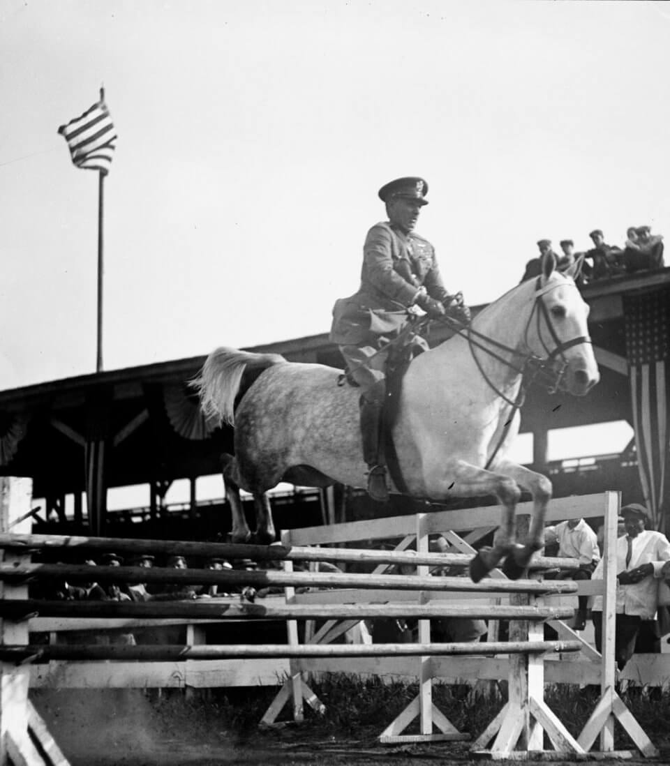 military rider jumping a fence with flag in the background
